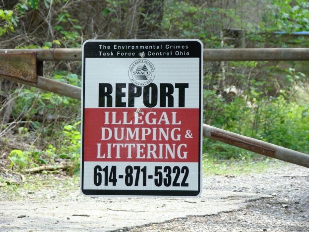 Sign on the roadside reading "Report Illegal Dumping and Littering" with trees and plants in the background and a board with text in the foreground.