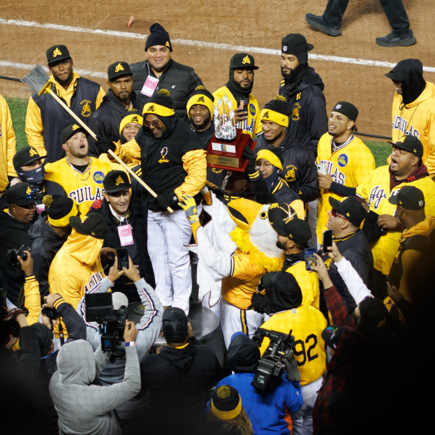 Gruppe von Baseball-Spielern in gelben und schwarzen Uniformen, die auf einem Feld feiern, einige halten einen Pokal und andere Kameras und Handys, mit sichtbaren Beinen im Hintergrund.
