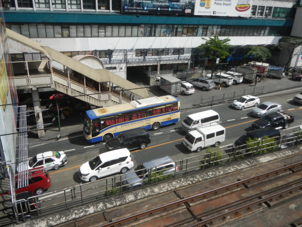 Eine belebte Stadtstraße mit Fahrzeugen, Gebäuden, Grünflächen und städtischer Infrastruktur, einschließlich Schildern und Bahnschienen.