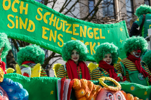 Eine Gruppe von Menschen in bunten Clownskostümen marschiert in einem Umzug, hält ein Banner, mit Bäumen und einem Gebäude im Hintergrund.