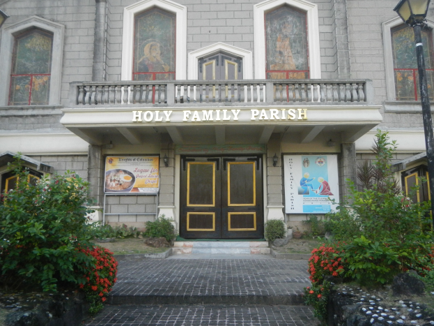 Die Heilige Familie Parish in Manila, Philippinen, mit einem Gebäude mit Fenstern, Türen, Geländern und einer Namensschild, Postern an den Wänden, Blumenpflanzen, Bäumen, einer Straßenlaterne und Steinen auf dem Boden unter einem bewölkten Himmel.