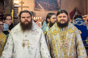 Zwei Priester in traditioneller russischer Orthodoxer Kleidung stehen Seite an Seite in einer Kirche, umgeben von Menschen in verschiedenen farbigen Kleidern, mit einem gerahmten Wandteil im Hintergrund und Gegenständen links.
