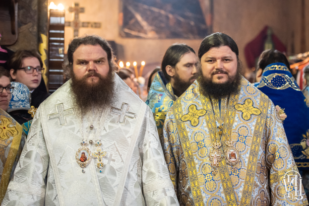 Zwei Priester in traditioneller russischer Orthodoxer Kleidung stehen Seite an Seite in einer Kirche, umgeben von Menschen in verschiedenen farbigen Kleidern, mit einem gerahmten Wandteil im Hintergrund und Gegenständen links.