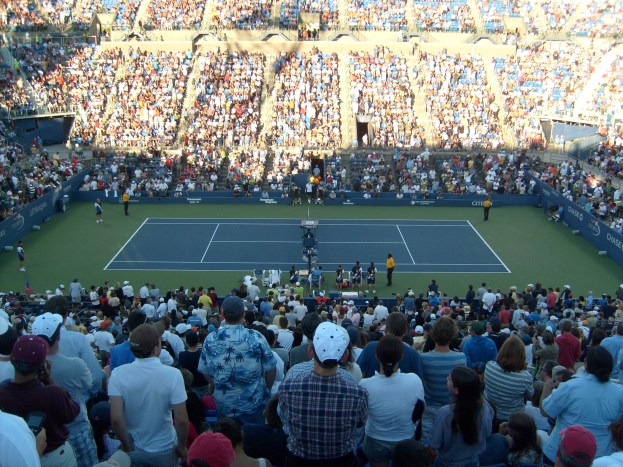 Eine große Zuschauermenge schaut einem Tennisspiel in einem vollen Stadion zu, mit Spielern auf dem Platz und Zuschauern in den Rängen.