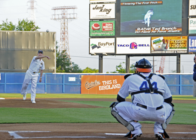 Ein Baseballspieler wirft einen Ball auf einem Feld mit einem Zaun, Werbetafeln, Strommasten mit Drähten, Bäumen und einem klaren blauen Himmel.