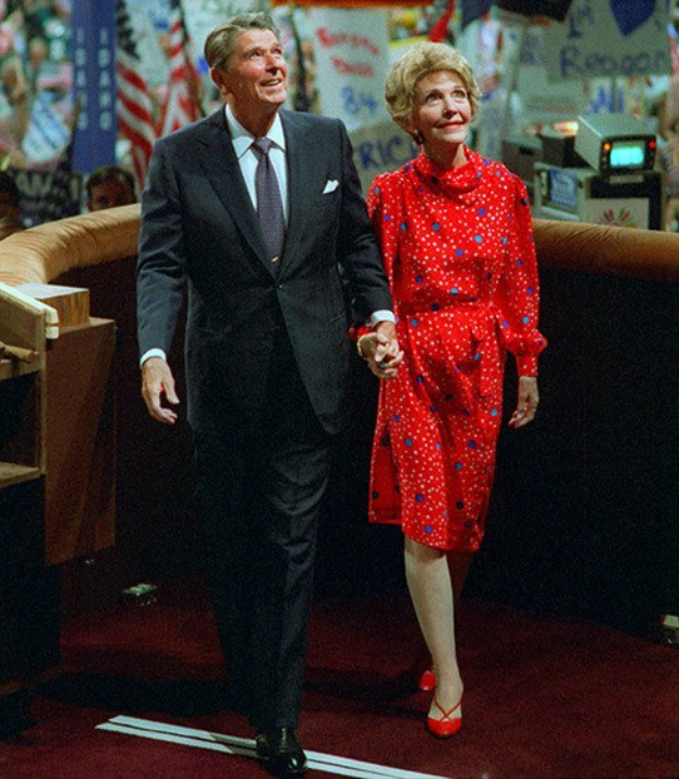Ein Mann in einem schwarzen Blazer, weißem Hemd und schwarzen Hosen sowie eine Frau in einem roten Kleid, die Hand in Hand eine rote Teppichbühne entlanggehen, mit Menschen, Fahnen, Bannern und anderen Gegenständen im Hintergrund.