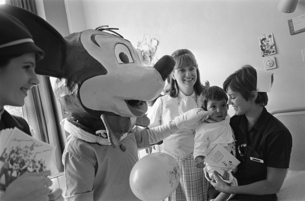 Eine Gruppe von Menschen, darunter eine Person in einem Mickey-Maus-Kostüm, versammelt sich um ein Baby im Krankenhausbett; zwei Frauen links halten Blumen und einen Ballon, mit gerahmten Bildern und einem Vorhang im Hintergrund.