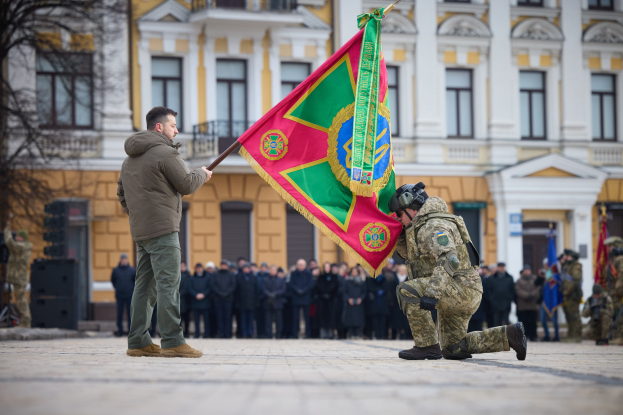 Ein Mann kniet neben einem anderen Mann, der eine Flagge hält, vor einer Menge, einige in Uniformen und Helmen, mit einem Gebäude und einem Baum im Hintergrund, wahrscheinlich während eines Militärparades zum 70. Jahrestag der Russischen Föderation.