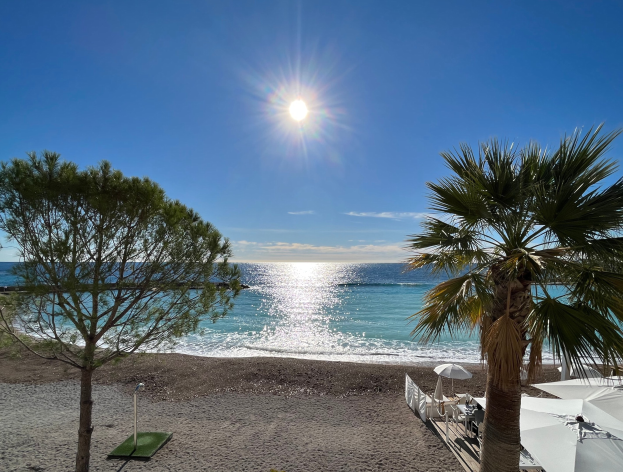 Eine sonnige Strandszene mit Palmen, Schirmen und üppiger Vegetation, vor einem strahlend blauen Himmel mit der Sonne im Hintergrund.