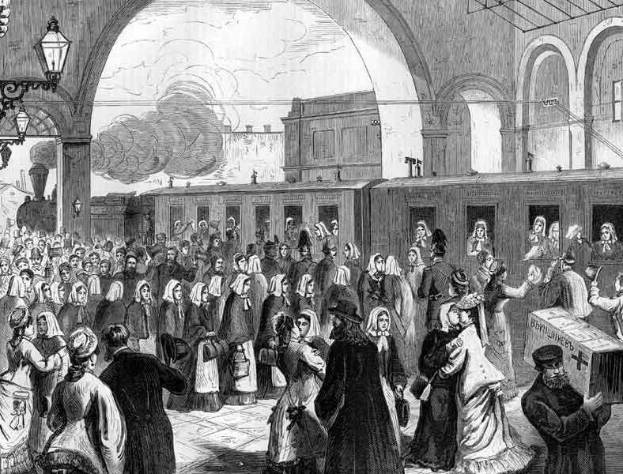 Schwarz-weißes Bild einer überfüllten Bahnhofshalle mit Menschen in verschiedener Kleidung, einem Zug auf den Schienen im Hintergrund, hängenden Lampen links und einer Person mit einer Kiste rechts, wahrscheinlich zeigt es die Ankunft von Flüchtlingen.