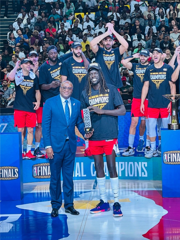 Gruppe von Männern auf einem Basketballfeld, die Trophäen halten, mit Zuschauern im Hintergrund und zwei Trophäen auf Podesten rechts.