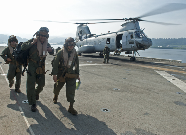 Menschen in Helmen und Schutzbrillen tragen Taschen und gehen auf das Flugdeck eines Flugzeugträgers zu, mit Wasser, Bäumen, Hügeln und einem klaren Himmel im Hintergrund.