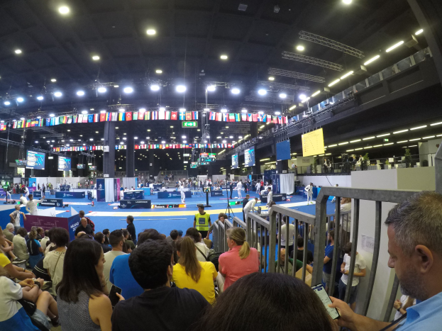 Eine große Menschenmenge auf Sitzen und Stehplätzen in einer Indoor-Sportarena mit Bildschirmen, Fahnen und Deckenleuchten im Hintergrund.