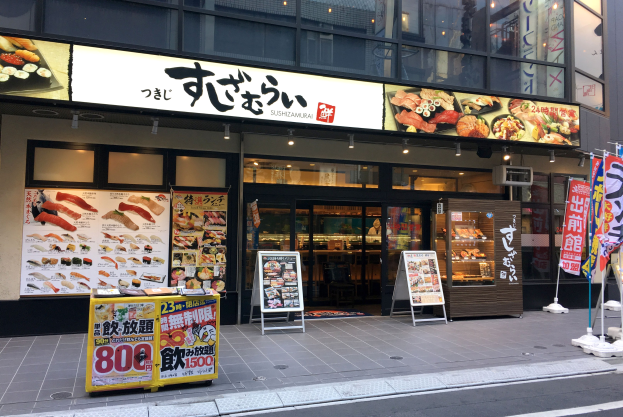 Außenansicht eines Sushi-Restaurants mit Glaswänden und -türen, das Speisekarten und beleuchtete Schilder zeigt, auf einer Tokioter Straßenkreuzung.