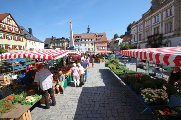 Ein belebter Markt im alten Stadtzentrum von Heidelberg, Deutschland, mit Menschen, die spazieren gehen, auf Bänken sitzen und in der Nähe von Zelten, Tischen mit Körben voller Gemüse und Gebäuden mit Fenstern, Bäumen und einem klaren blauen Himmel im Hintergrund.