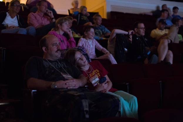 Eine Gruppe von Männern und Frauen sitzt in einem Kinosaal mit einem Bildschirm auf der rechten Seite und einer Wand im Hintergrund.