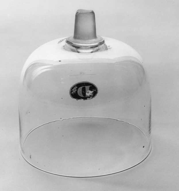 Schwarz-weißes Foto eines Glasglashauses mit Metalldeckel, zylindrisch mit breiter Basis und engem Hals, Deckel leicht angehoben, enthüllt Inhalt.