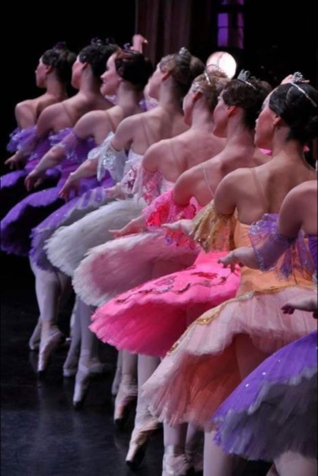 Eine Gruppe von Balletttänzerinnen in rosafarbenen Tutus sitzt auf einer Bühne, beleuchtet von einem Licht im Hintergrund, und bewegt sich synchron.