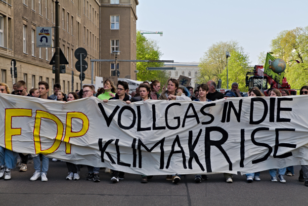 Group of people walking down a street holding a banner reading "FDP Vollgas in die Klimakrise" with buildings, trees, and traffic signals in the background under a clear blue sky.