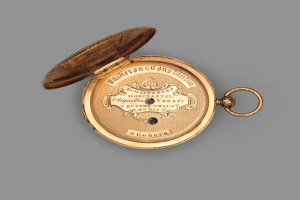 Goldene Taschenuhr mit geschlossenem Deckel auf grauem Hintergrund, mit eingravierter Inschrift, die wahrscheinlich die Marke oder Art angibt.