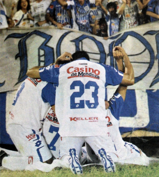 Eine Gruppe von Fußballspielern feiert auf dem Platz vor einer jubelnden Menge, im Hintergrund ist eine Fahne mit Text zu sehen.