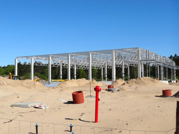 Eine Baustelle mit einer großen Metallstruktur in der Mitte, umgeben von Sand, Pfählen, einem Zaun und anderen Gegenständen, mit Bäumen und einem klaren blauen Himmel im Hintergrund.