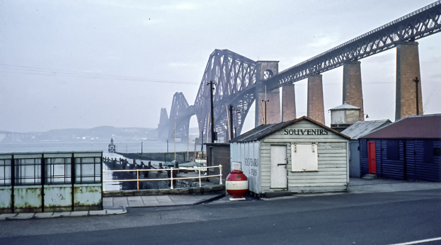 Die Forth-Eisenbahnbrücke in Schottland überspannt Wasser mit Hügeln und Wolken im Hintergrund, während eine Straße, Schuppen, Pfähle und ein Geländer im Vordergrund zu sehen sind.