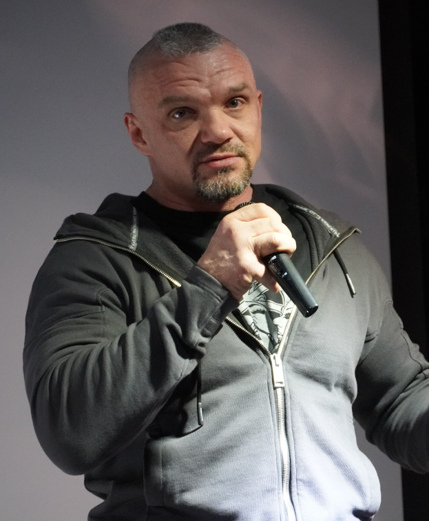 Ein Mann in einer schwarzen Kapuzenjacke und einem schwarzen Anzug mit einem weißen Hemd, glatt zurückgekämmtem Haar und einem selbstbewussten Gesichtsausdruck hält ein Mikrofon, während er vor einer Wand steht.