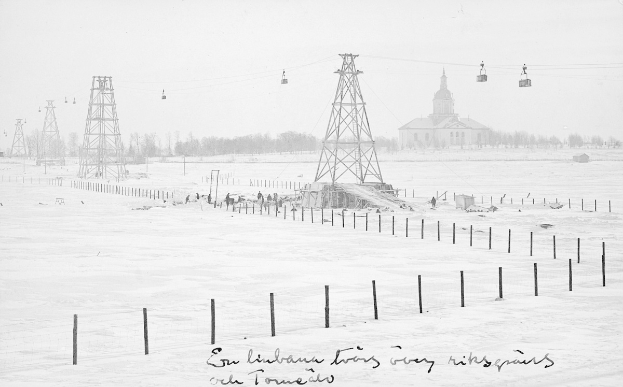 Schwarze-Weiß-Foto eines Skilifts in einem verschneiten Feld mit Stützpfählen, Überseilbahn, Bäumen und einem Gebäude im Hintergrund; Text unten.