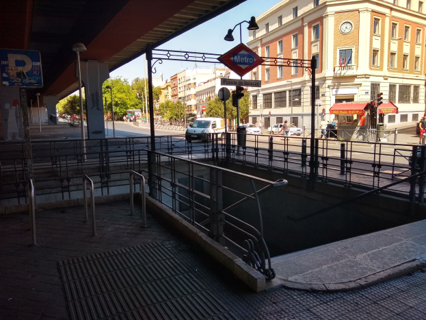 Eine belebte Stadtstraße mit Fahrzeugen, Fußgängern, Gebäuden, einer Brücke mit Geländern, einem Schild, Bäumen, einem klaren blauen Himmel und einem zentralen Springbrunnen.