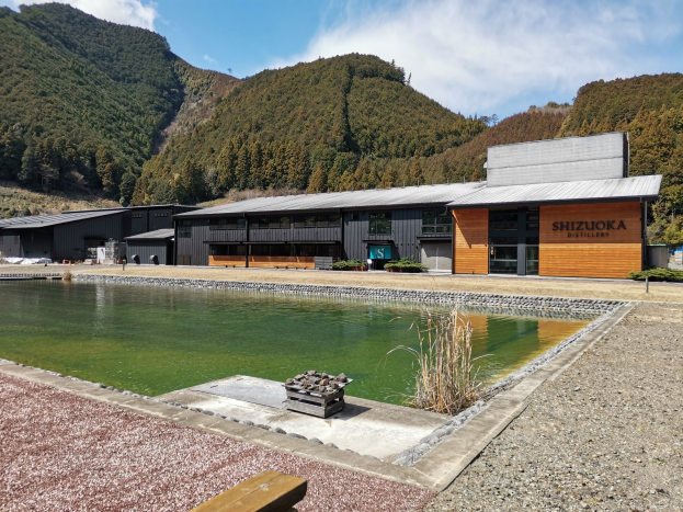 Großes cremefarbenes Gebäude mit einer Namenstafel, das vor grünen Hügeln unter einem bewölkten Himmel steht und einen Teich im Vordergrund hat, der von Bäumen und Pflanzen umgeben ist.