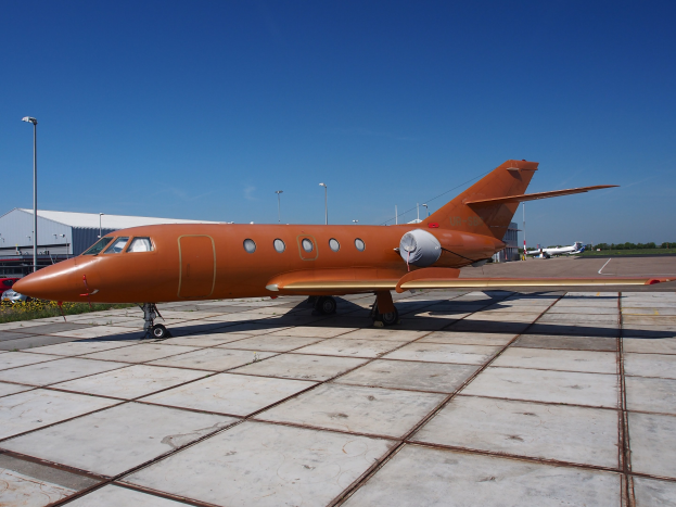 Ein privater Jet auf einem Flughafenvorfeld mit umgebenen Fahrzeugen, Gebäuden, Polen, Lampen, Pflanzen und Bäumen unter einem klaren blauen Himmel.