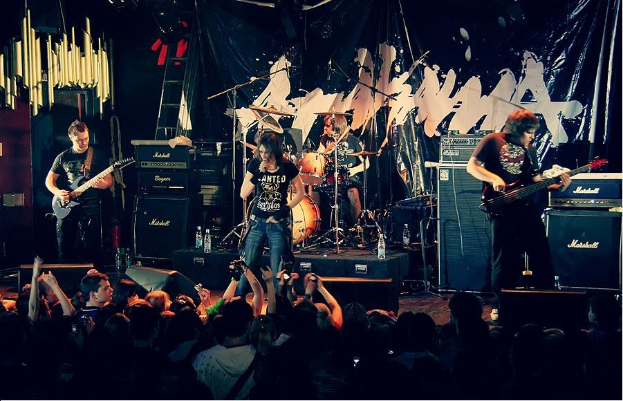 Eine Gruppe von Musikern, die auf der Bühne mit Gitarren und Trommeln, Mikrofonen und einem Banner im Hintergrund spielen, mit Text unten, der "The Wanted - Live at the O2 Academy, London" liest.