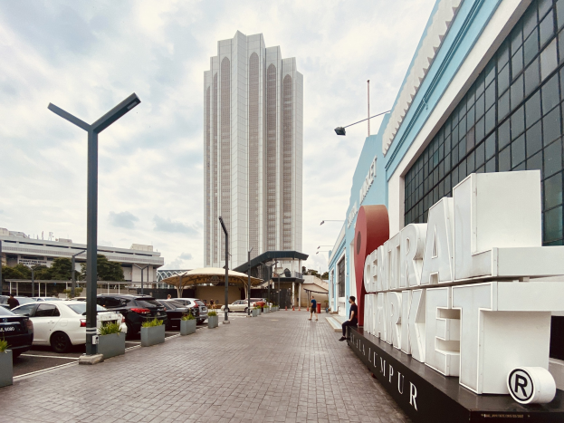 Ein belebter zentraler Markt in Kuala Lumpur, Malaysia, mit hohen Gebäuden, Straßenlaternen, Straßenschildern, Motorfahrzeugen, Fußgängern, Namenstafeln, Bäumen und einem bewölkten Himmel.