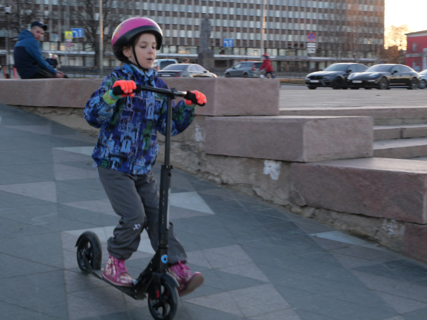 Ein junger Junge mit Helm und Handschuhen fährt auf einem Roller eine Treppe hinunter, vorbei an Fahrzeugen, Menschen, Bäumen, Pfosten, Brettern, Gebäuden und einem klaren blauen Himmel.
