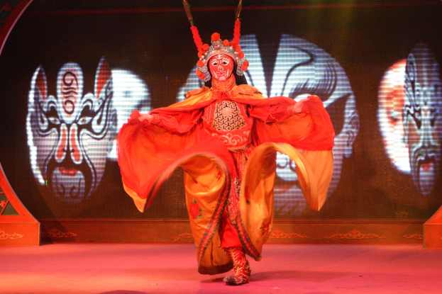 Eine Frau in einem roten Kleid führt eine chinesische Oper auf der Bühne auf, trägt ein farbenfrohes Kostüm und bewegt sich anmutig zur Musik, mit einem Sichtschutz im Hintergrund.