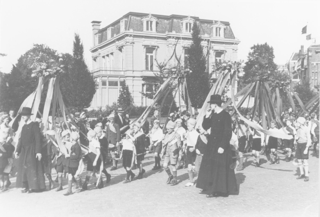 Schwarze und weiße Aufnahme einer Gruppe von Menschen, die eine Straße entlanggehen und Fahnen tragen, mit Hüten, Bäumen, Gebäuden und einem Maibaum im Hintergrund.