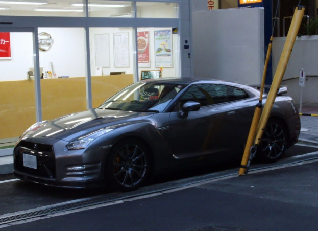 Ein schwarzer Nissan GT-R vor einem Gebäude mit einer Stange auf der rechten Straßenseite, Schilder mit Text und Bildern im Hintergrund und Deckenbeleuchtung.