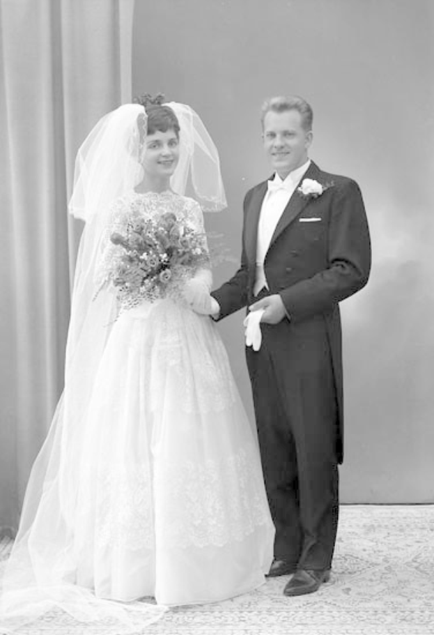 Schwarzes und wei├čes Foto eines l├Ąchelnden Mannes und einer Frau, die nebeneinander auf ihrer Hochzeit stehen, die Frau in einem wei├čen Kleid mit einem Strau├č, mit einem Vorhang und einer Wand im Hintergrund.