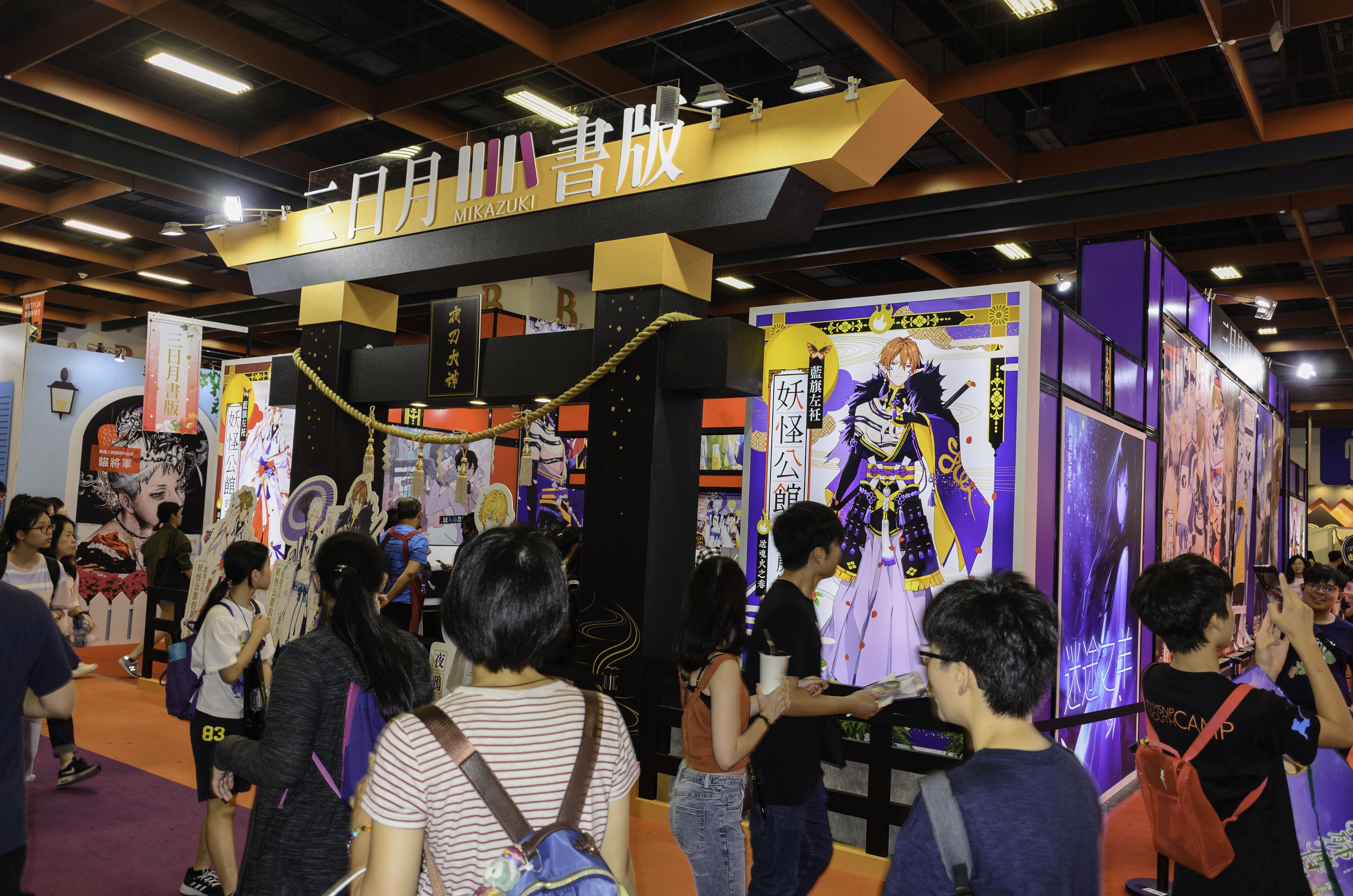 Gruppe von Menschen an einem Anime-Convention-Stand in Tokio mit Bannern an den Wänden, einer Kettenbarriere und Deckenbeleuchtung.