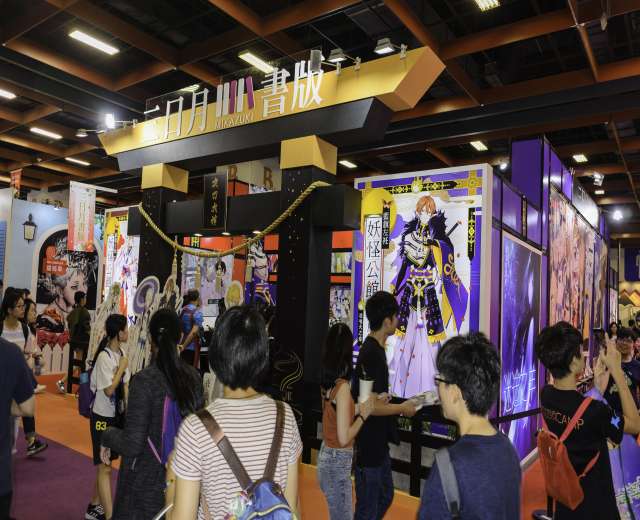 Gruppe von Menschen an einem Anime-Convention-Stand in Tokio mit Bannern an den Wänden, einer Kettenbarriere und Deckenbeleuchtung.