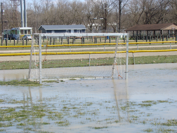 Fußballtor in einem überfluteten Feld mit Schuppen, Pfählen, Bäumen und Fahrzeugen im Hintergrund unter einem klaren blauen Himmel.