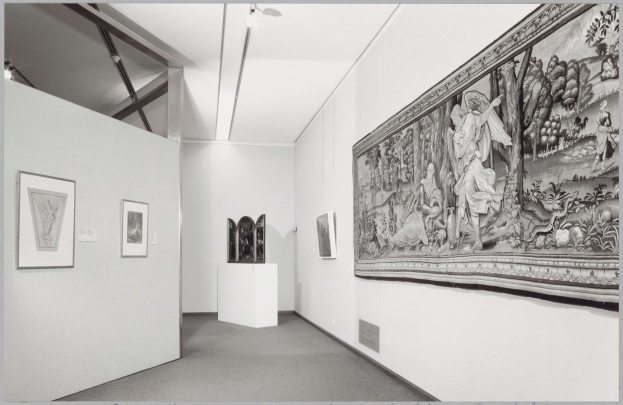 Schwarzes und weißes Foto eines Museumsraums mit einem großen Wandteppich, gerahmten Kunstwerken an den Wänden, Sockelobjekten, Deckenleuchten und "Museum of Fine Arts, New York City"-Text unten.