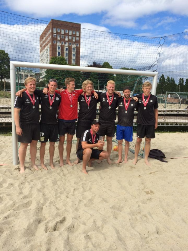 Mannergruppe in Sportkleidung auf einem sandigen Strand stehend, mit Medaillen und einem Netz umgeben, mit Bäumen, Gebäuden und einem bewölkten Himmel im Hintergrund.
