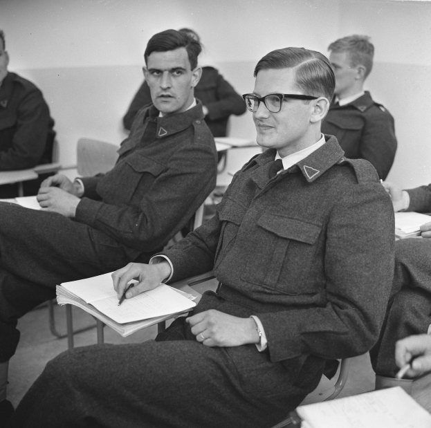Schwarzes und weißes Foto von Männern in Militäruniformen, die in einem Klassenzimmer sitzen und Bücher und Stifte halten.