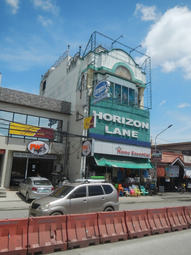 Eine belebte Straßenszene mit Fahrzeugen, Absperrungen, Gebäuden einschließlich des Horizon Lane Hotels in Manila, elektrischen Polen mit Drähten und einem bewölkten Himmel.
