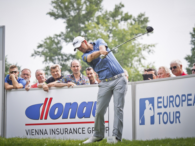 Ein Golfer in einer Mütze schwingt einen Schläger auf einem grünen Feld mit Sponsor-Wänden, Bäumen und einem blauen Himmel im Hintergrund, was das Ende der European Tour anzeigt.