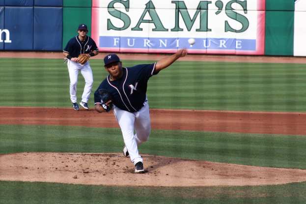 Baseball-Spieler wirft auf einem grünen Feld mit zwei Personen in Mützen und Handschuhen im Hintergrund und einem Banner mit Text.
