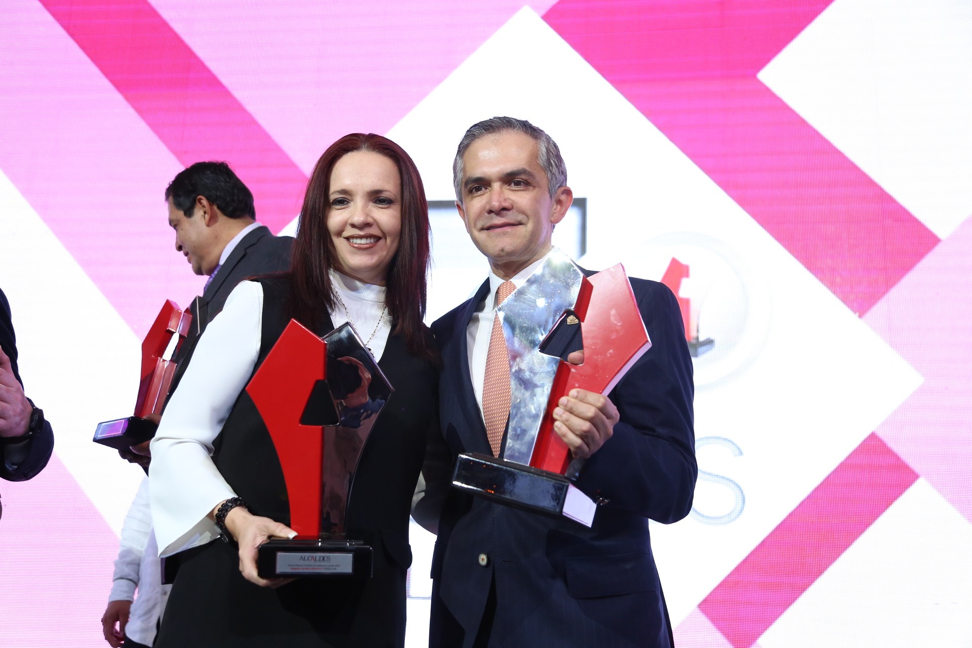 Zwei Personen auf einer Bühne mit Trophäen stehend und strahlend, mit einem Awards-Ceremony-Bildschirm im Hintergrund.