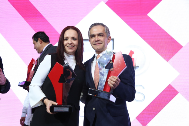 Zwei Personen auf einer Bühne mit Trophäen stehend und strahlend, mit einem Awards-Ceremony-Bildschirm im Hintergrund.